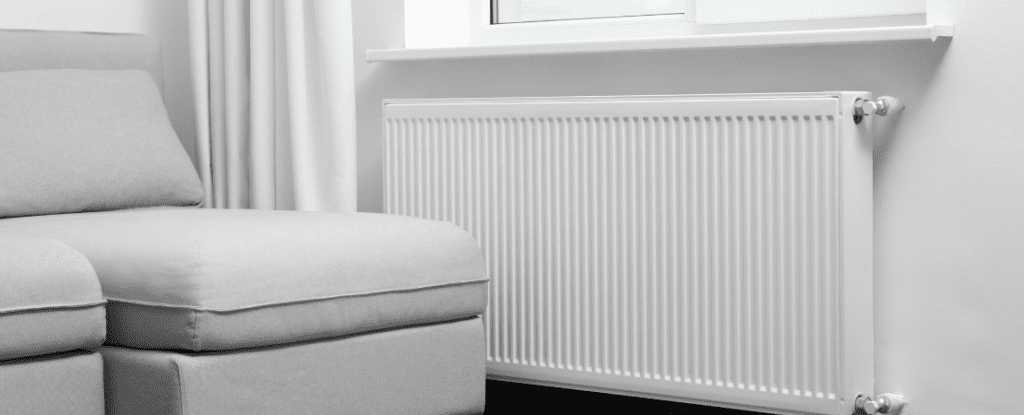 Moderner Niedrigtemperatur-Heizkörper unter einem Fenster in einem hell eingerichteten Wohnzimmer. Daneben steht ein graues Sofa mit Stoffbezug, eine weiße Gardine hängt im Hintergrund. Symbolbild für energieeffizientes Heizen, moderne Heiztechnik und Wohnkomfort.